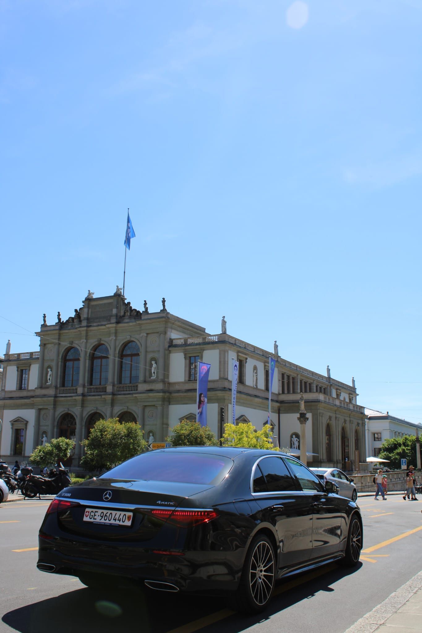 Une berline noire passe devant un bâtiment historique en pierre sous un ciel d'un bleu éclatant.
