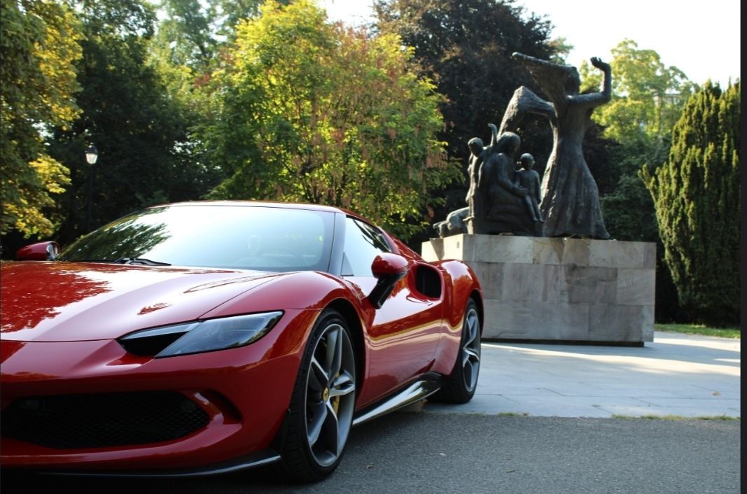 Une voiture de sport rouge est garée à l'extérieur, à côté d'une statue sombre sur un piédestal en pierre, entourée d'arbres.