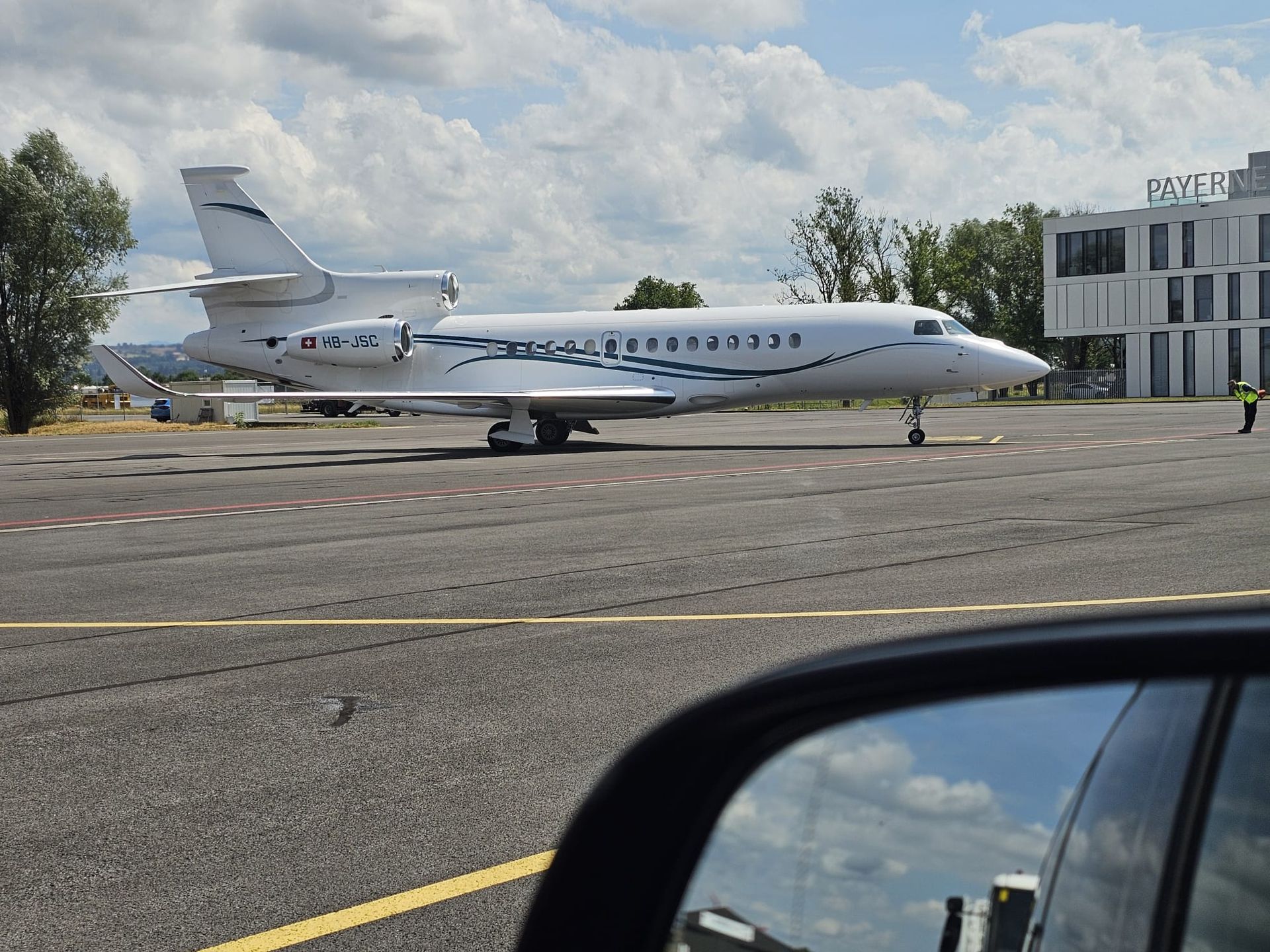 Un jet d'affaires blanc sur le tarmac d'un aéroport, près d'un bâtiment, vu depuis le rétroviseur d'une voiture.