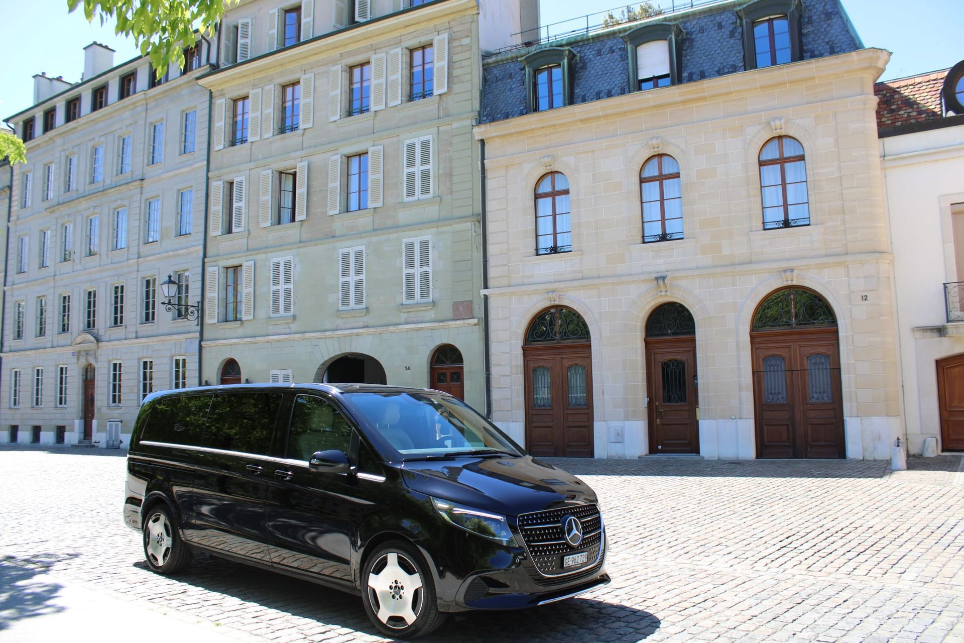 Une camionnette noire garée dans une rue pavée ensoleillée, à côté de bâtiments en pierre claire aux portes et fenêtres cintrées.