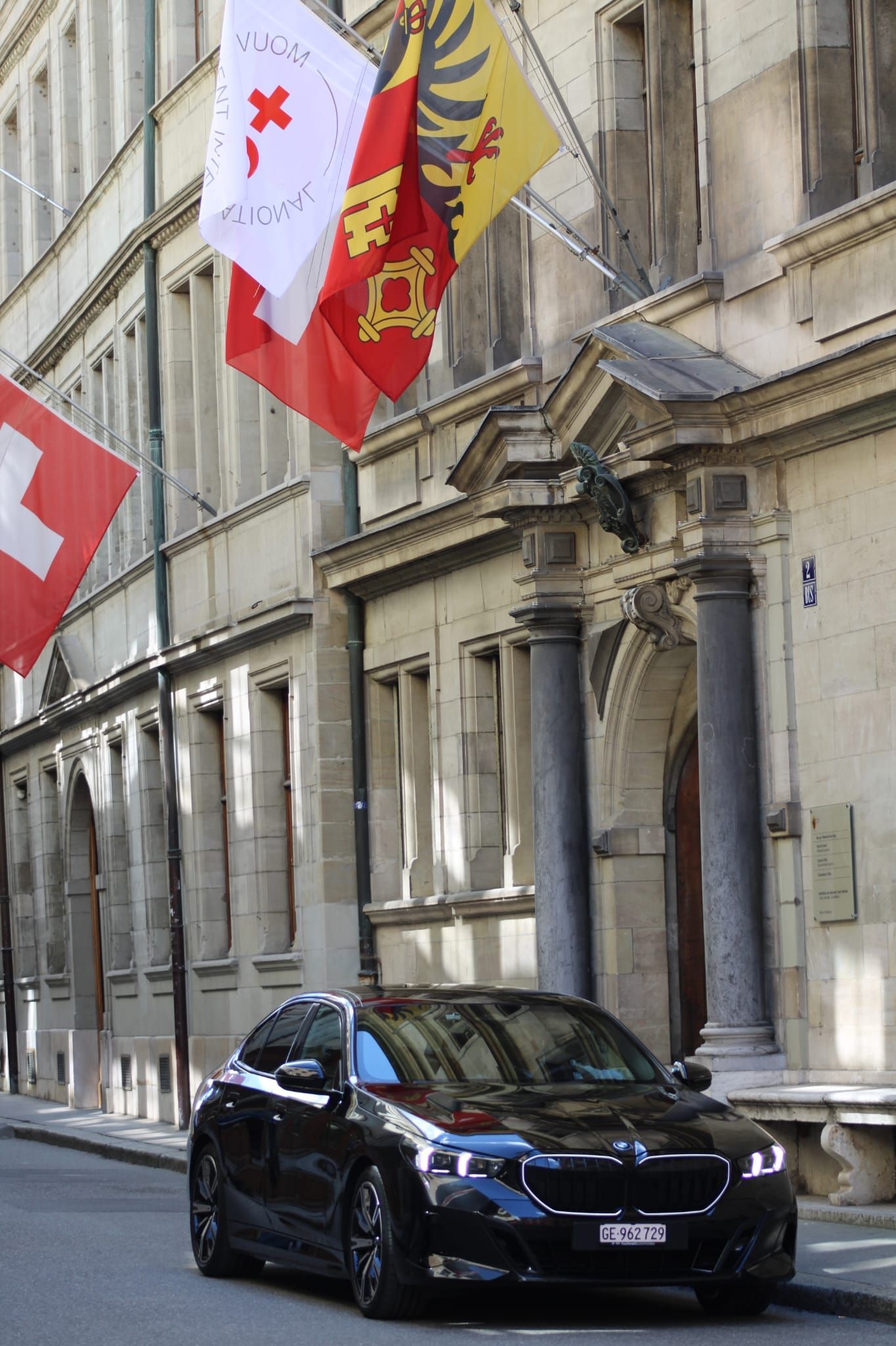 Voiture noire garée dans une rue étroite, à côté d'un bâtiment en pierre orné de drapeaux rouges et blancs.