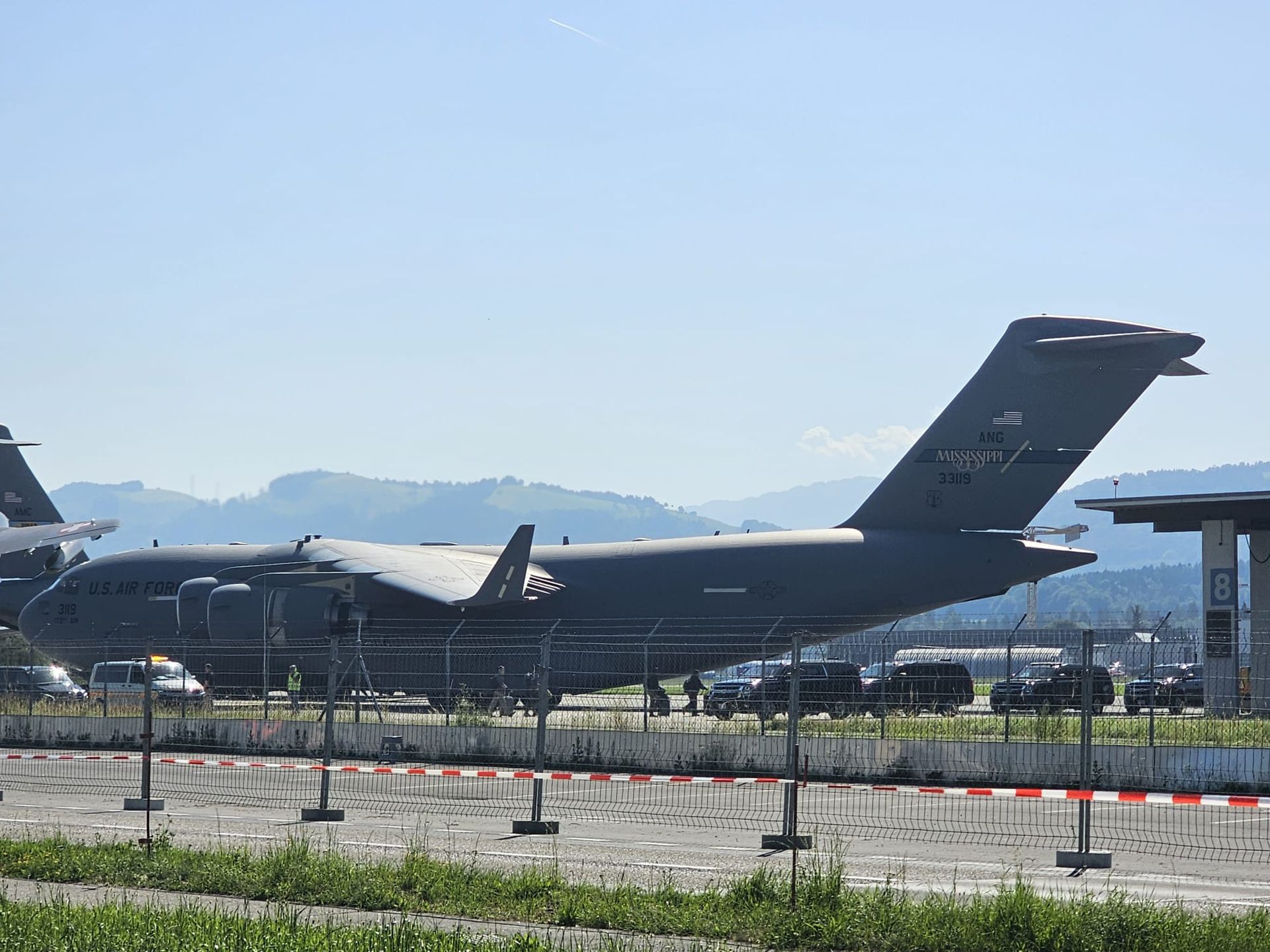 Avion cargo militaire stationné sur une piste, vu derrière une clôture avec des montagnes en arrière-plan.