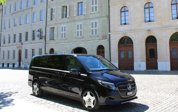 Une camionnette noire garée dans une rue pavée ensoleillée, à côté d'un bâtiment beige aux fenêtres cintrées.