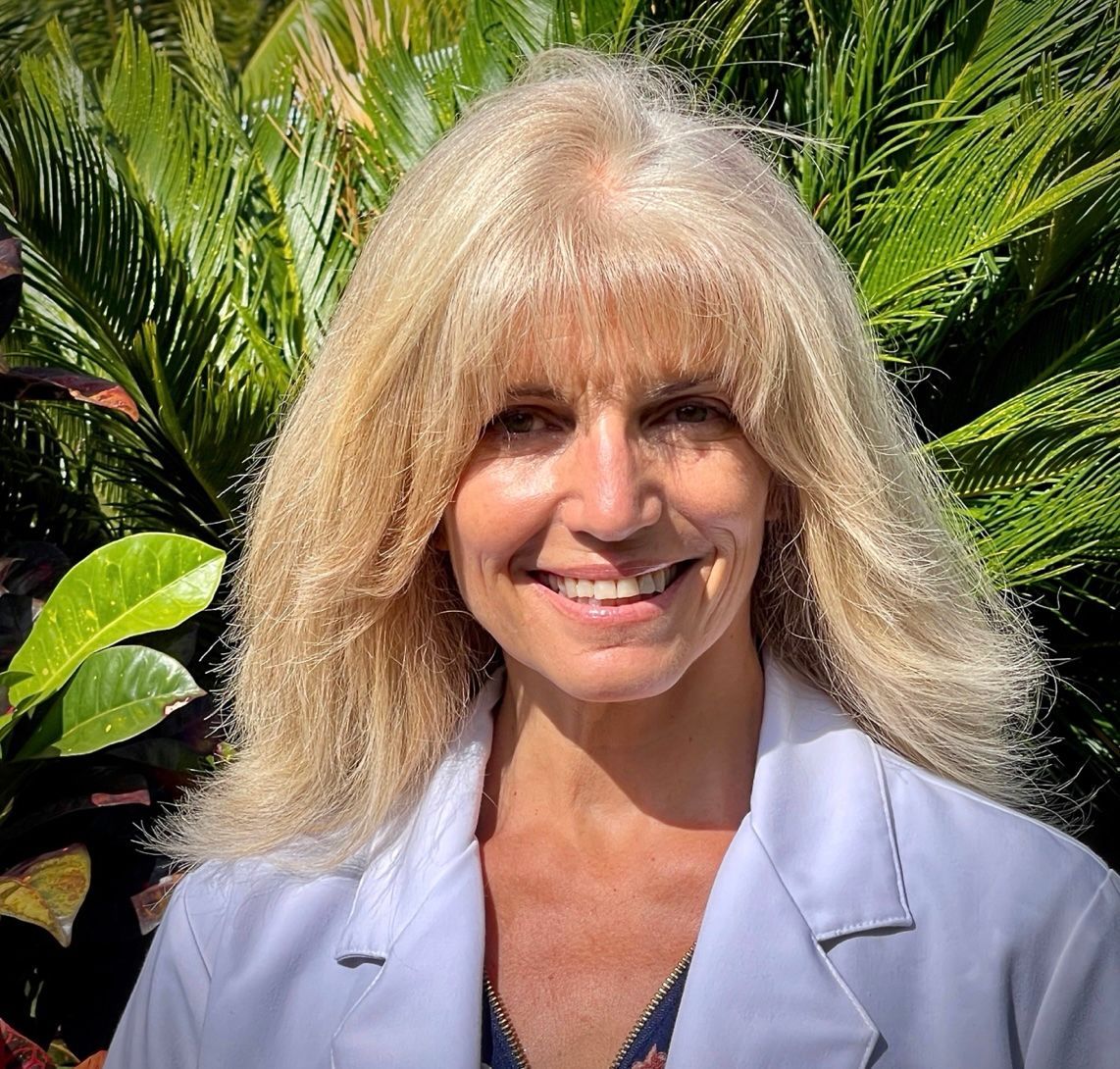 Smiling blonde woman in a white lab coat in front of lush green plants.