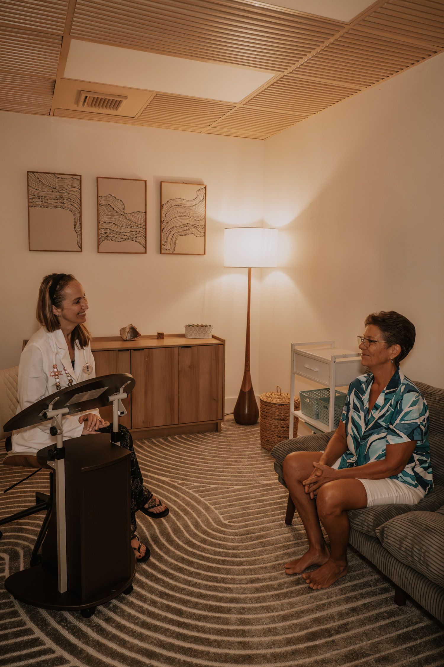 Doctor and patient in a calming room. The doctor listens to the patient, who is sitting on a sofa. The room has wood accents.