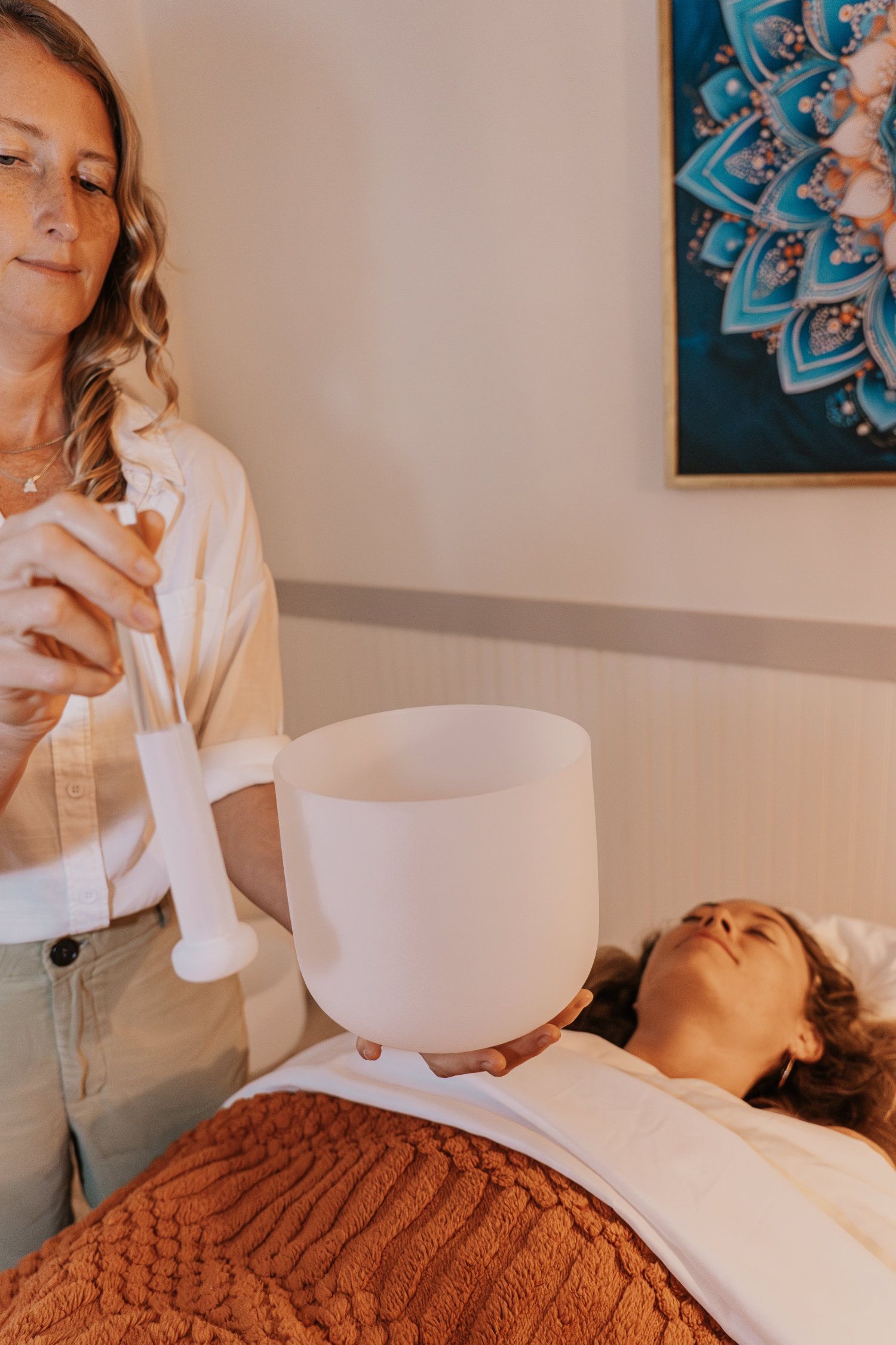 Woman holding a crystal bowl over another woman, offering sound healing in a room.