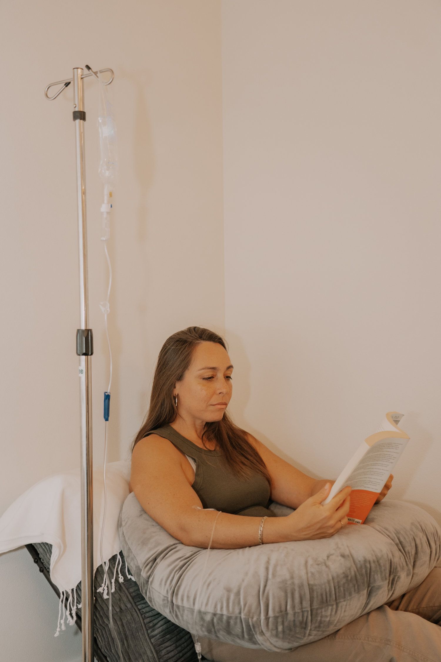 Woman reading while receiving an IV infusion in a room, sitting on a pillow with a medical stand nearby.