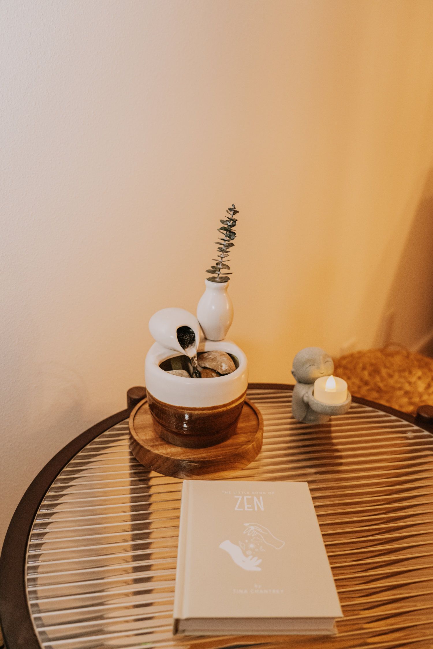 A small fountain with greenery on a round table, a book, and a statue. Soft lighting, neutral colors.
