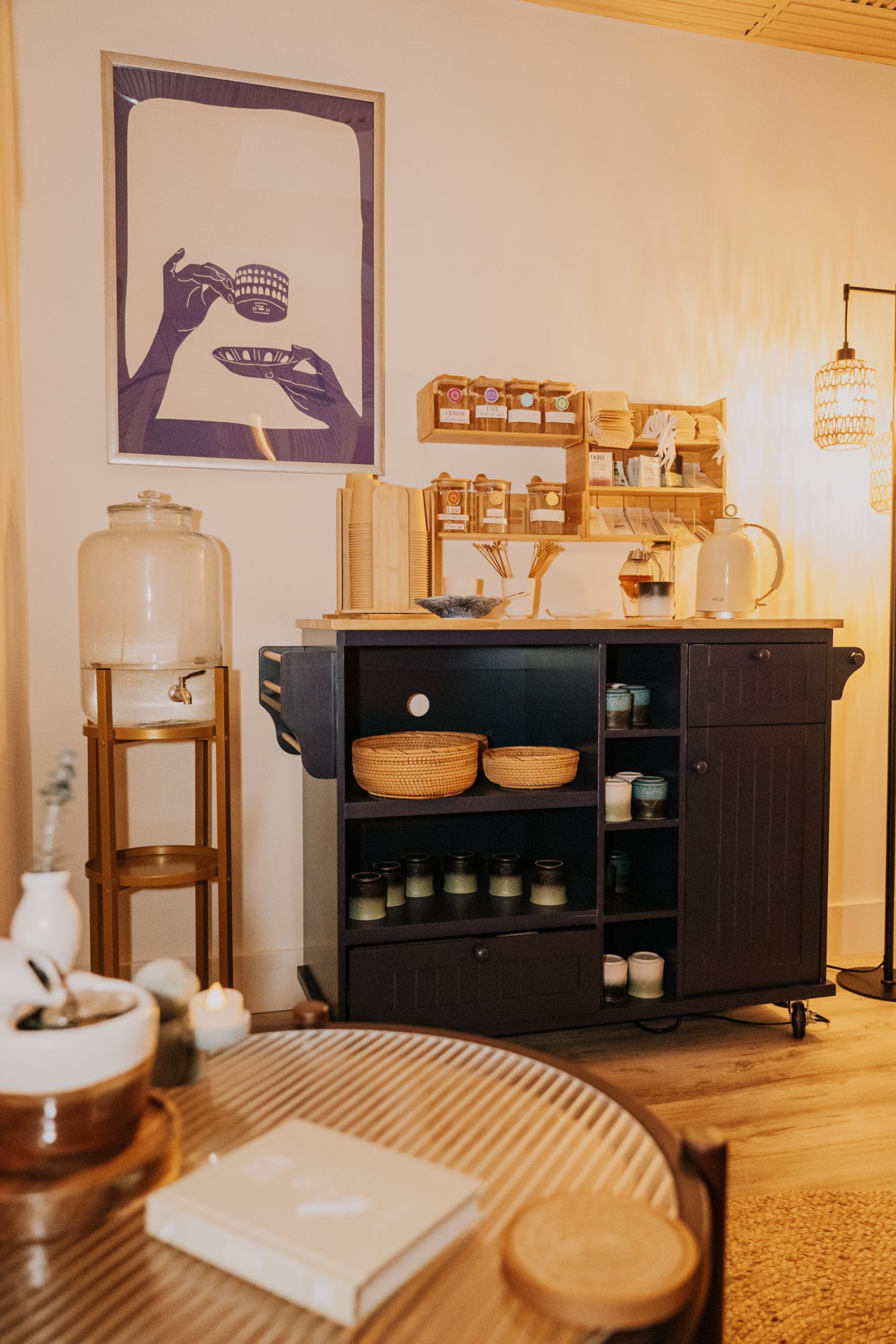 A dark cabinet tea station with shelves of jars, a water dispenser, and a round table.