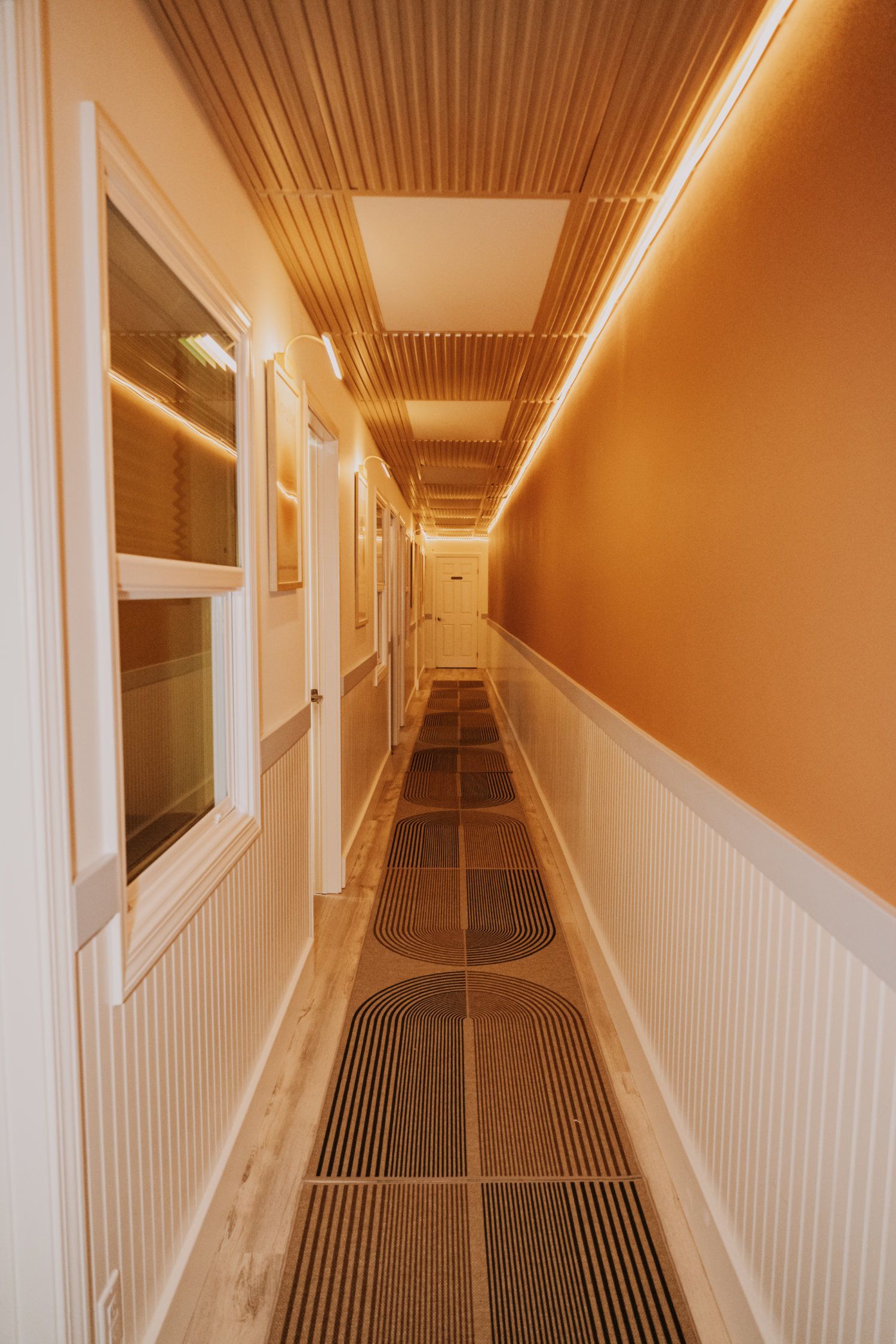 Long hallway with patterned floor, white paneling, orange wall, and ceiling lighting.