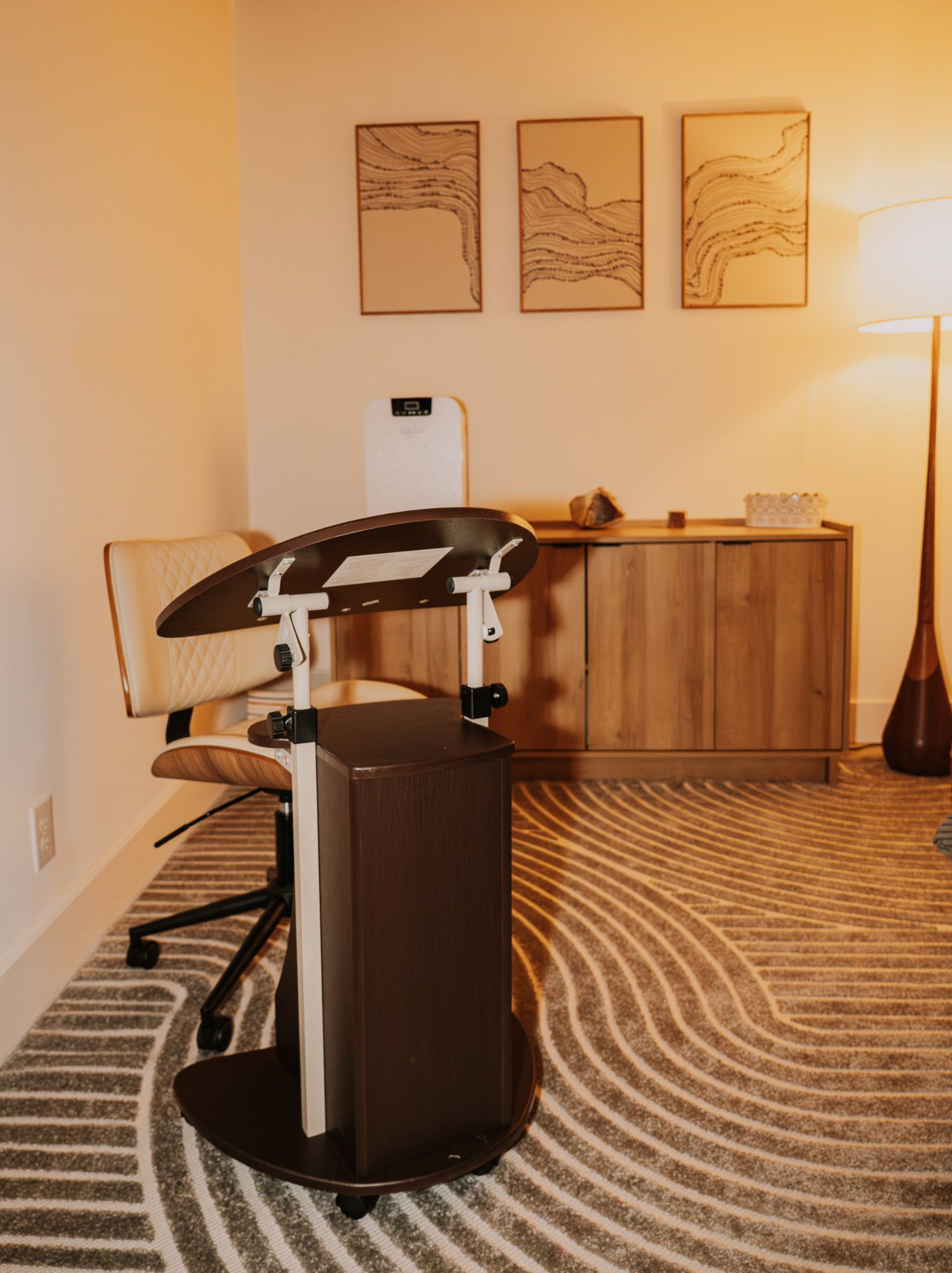A rolling office workstation in a room with a rug, desk, chair, artwork, and a floor lamp.
