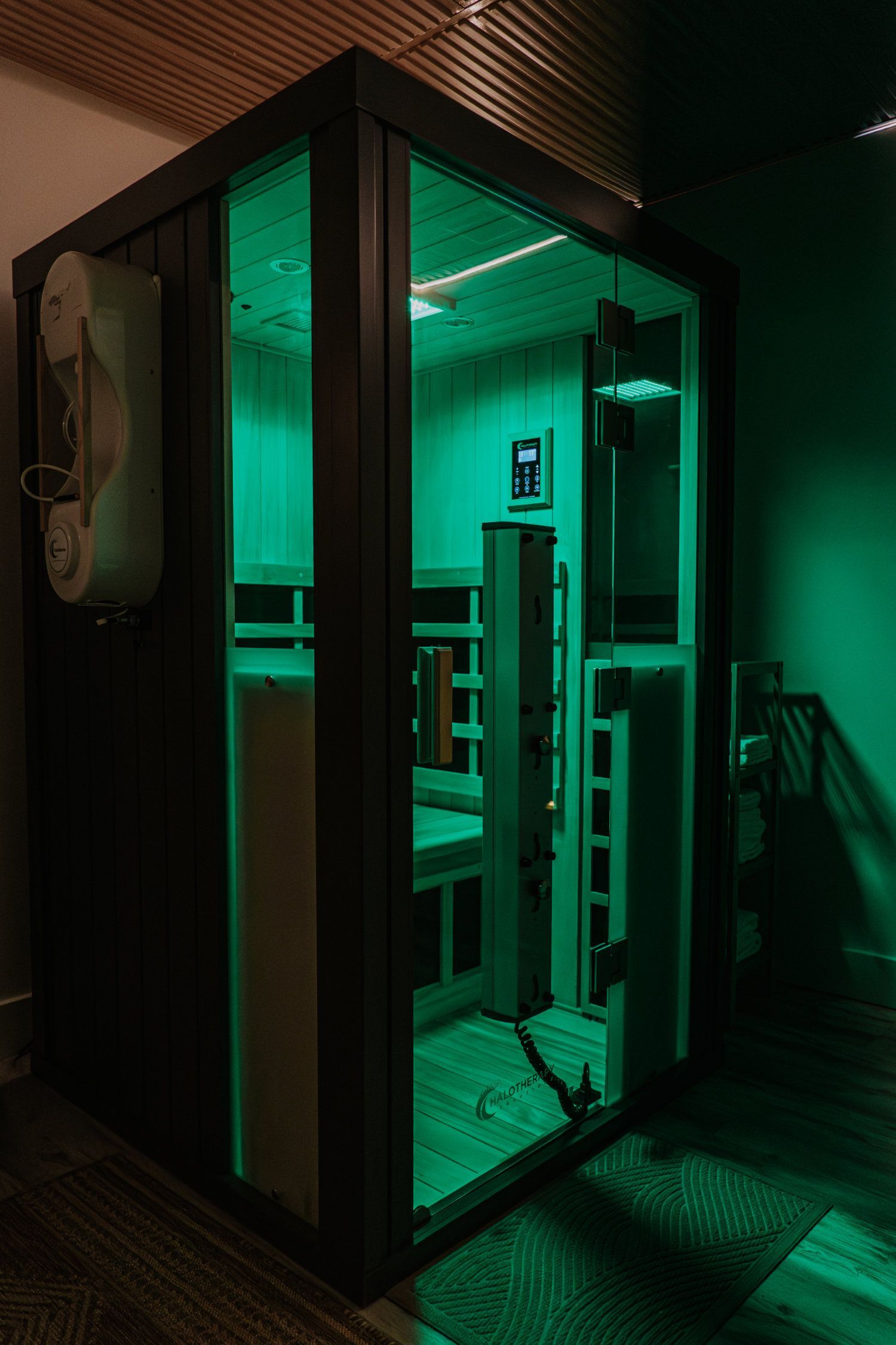 A dark wood infrared sauna with green interior lighting. A control panel is visible inside the glass door.