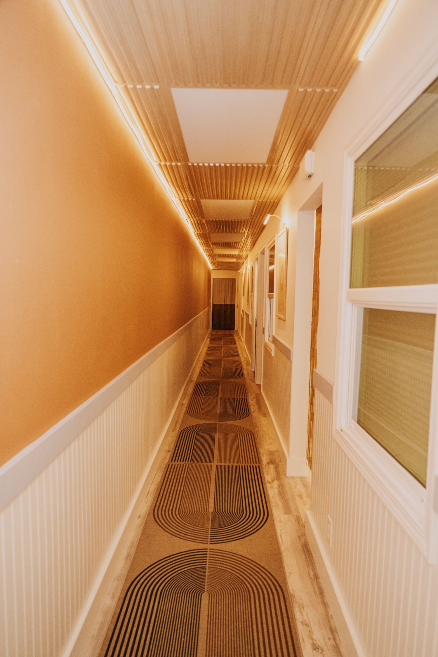 Long hallway with patterned floor, peach wall, and wood-paneled ceiling. Windows line one side.