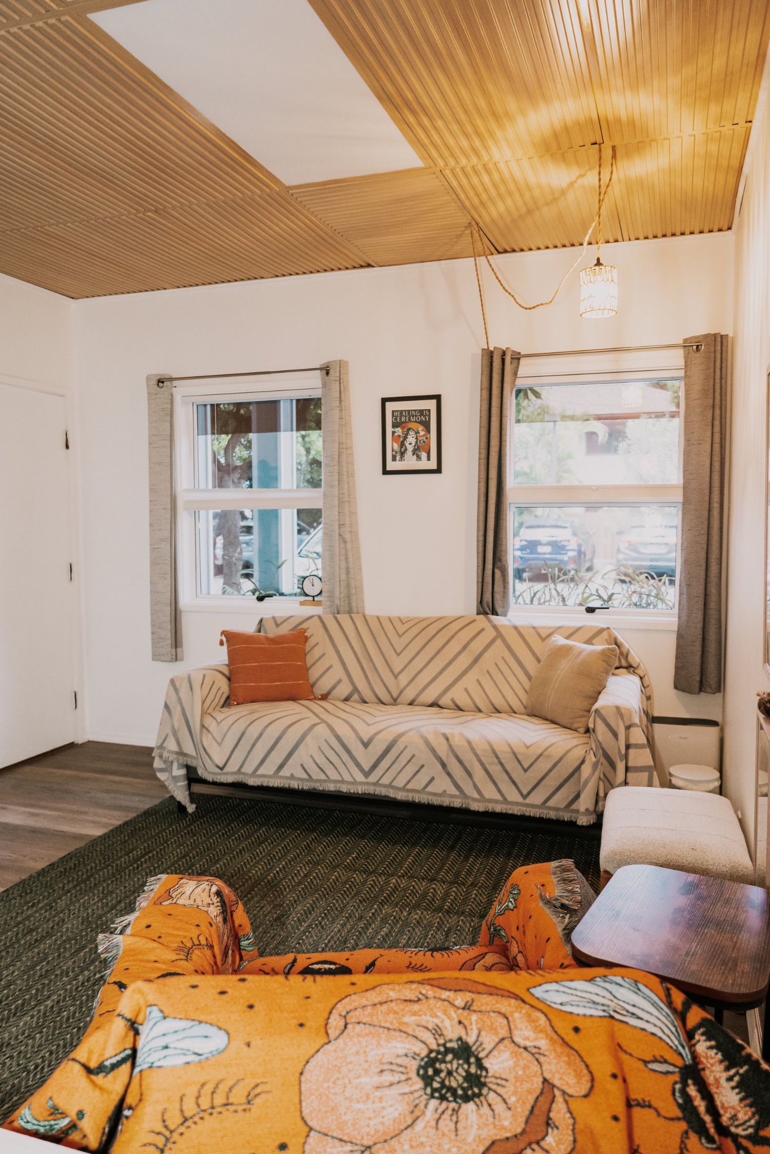 Cozy room with a sofa, windows, green rug, and a patterned blanket, under a woven ceiling.