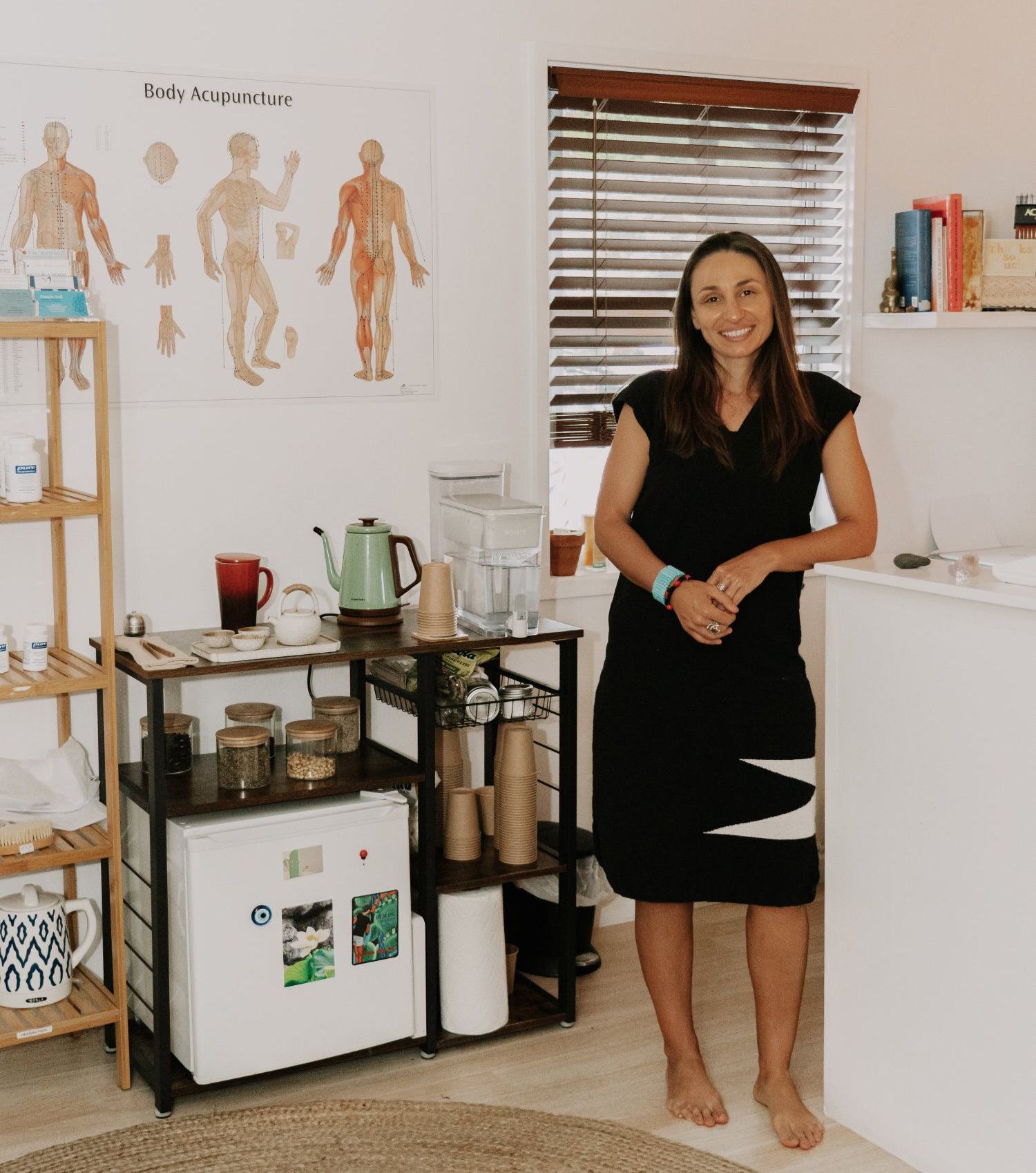 Woman in black dress, smiles, stands in her treatment room, near rolling cart with supplies.