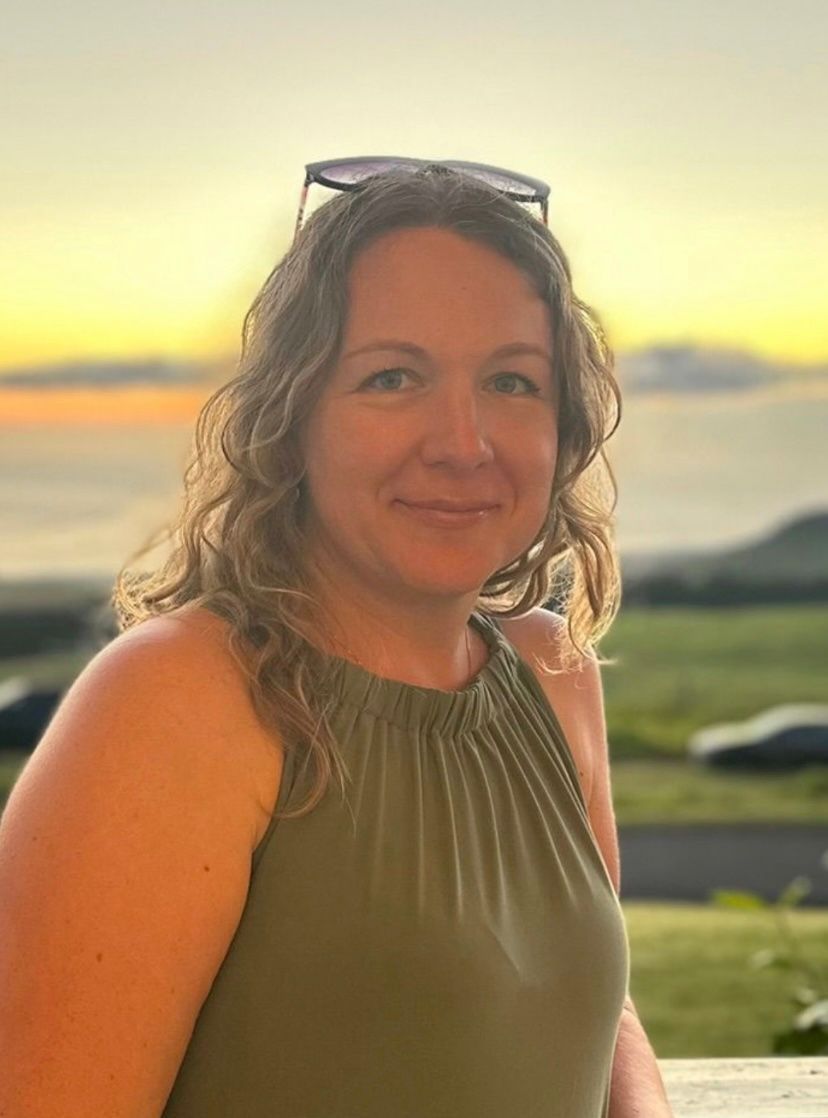 Woman with blonde hair, smiling, wearing a black top and gold jewelry, in front of a light blue wall.