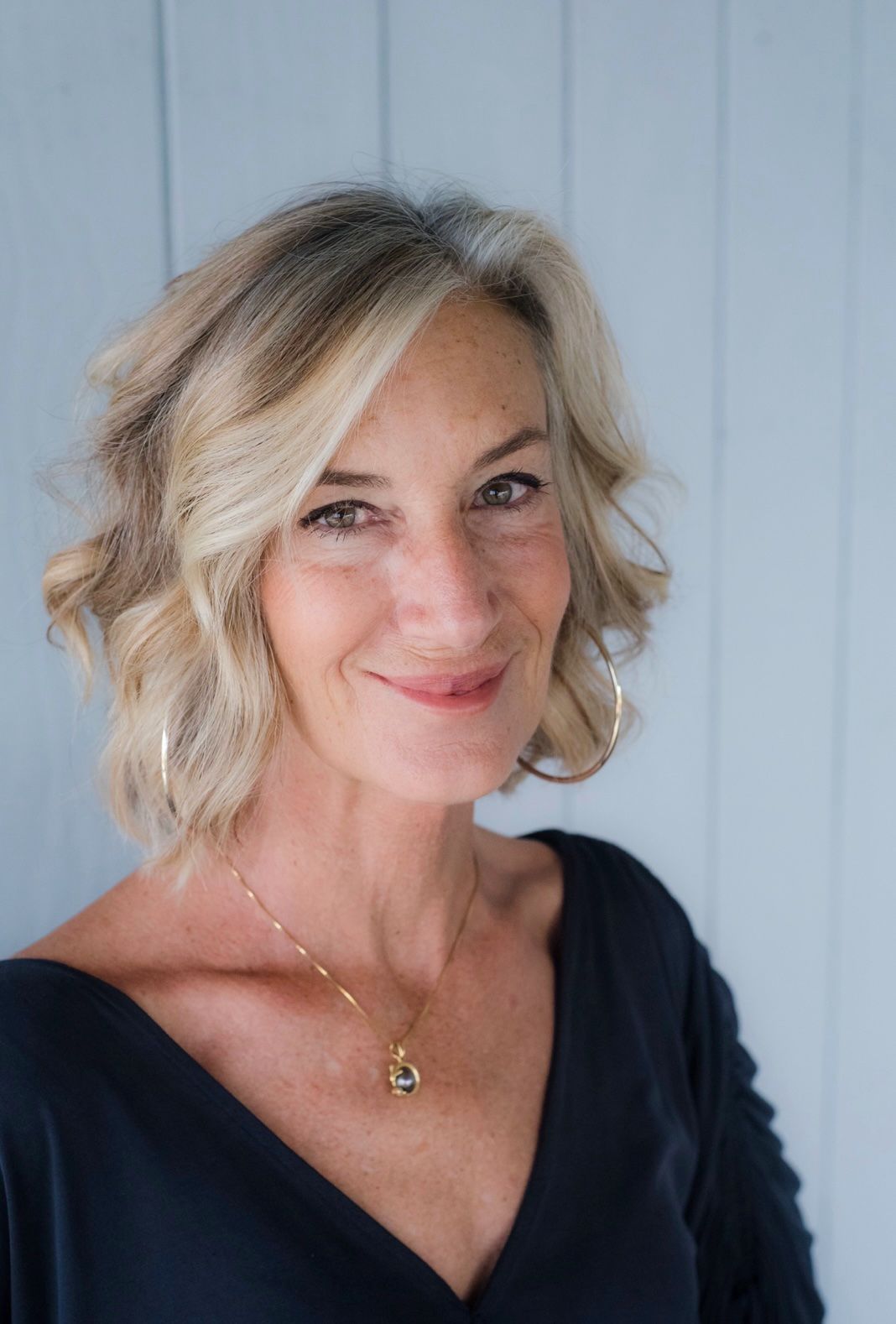Woman with blonde hair, smiling, wearing a black top and gold jewelry, in front of a light blue wall.