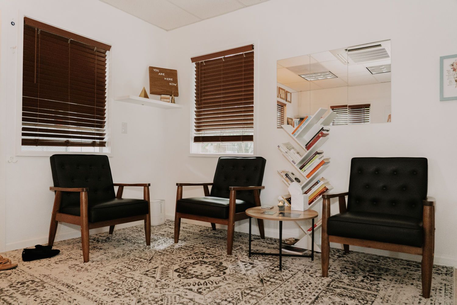 Three black chairs in a waiting room with brown blinds, a mirror, and a small table on a patterned rug.