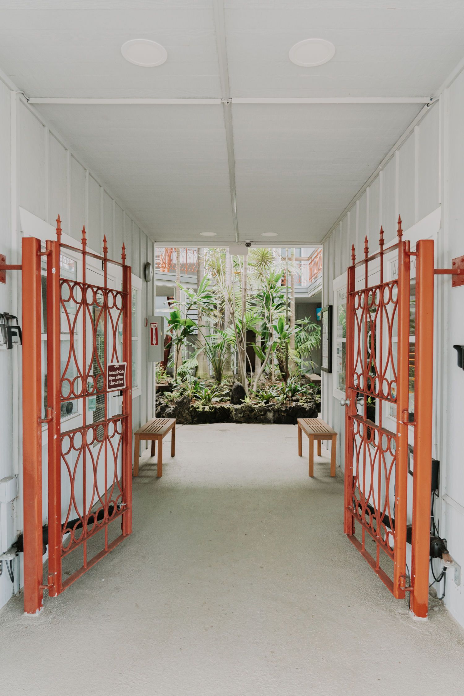 Open red gates lead to a garden. White hallway, benches, and foliage in the distance.