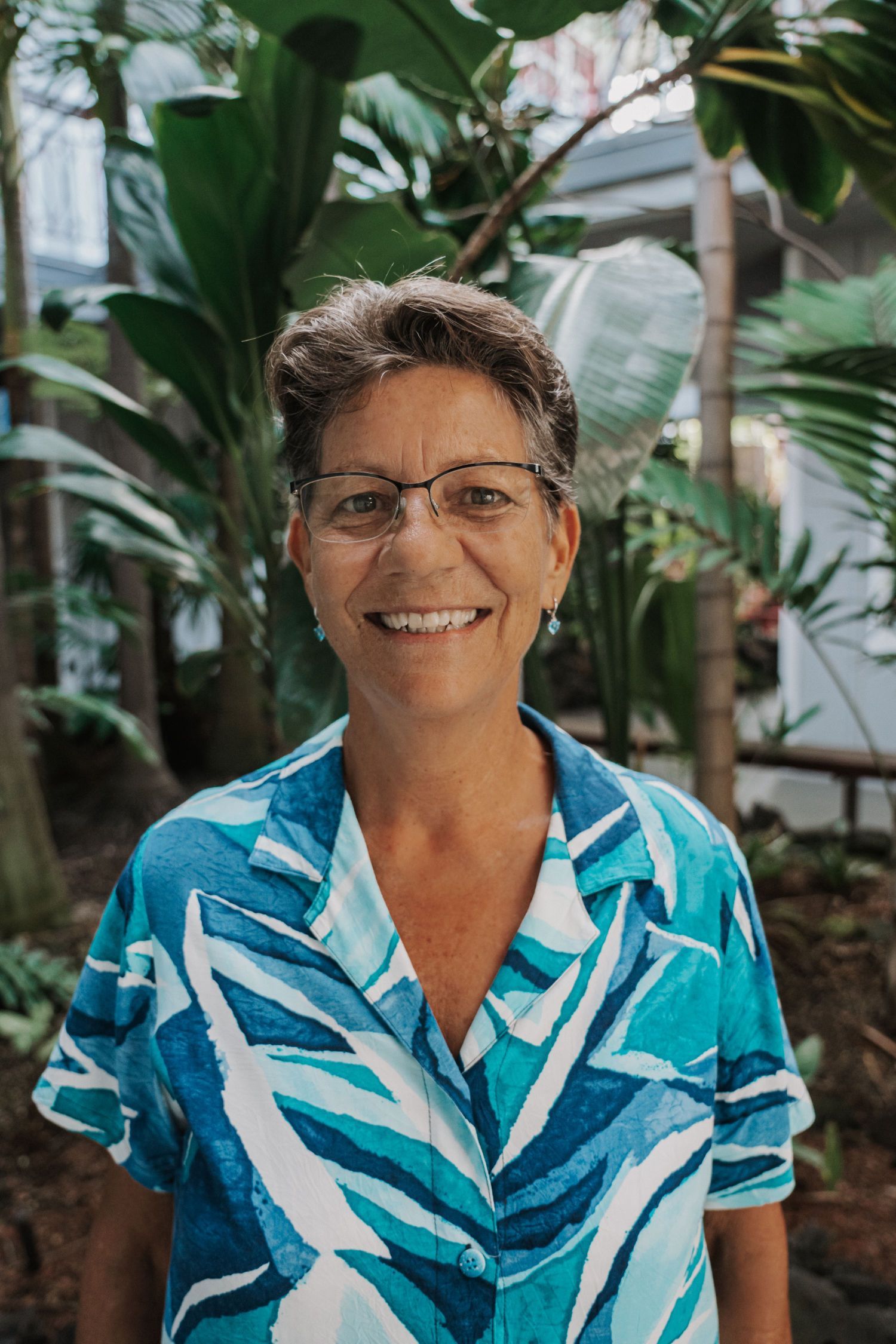 Woman with glasses smiles, wearing a blue patterned shirt, outdoors in front of tropical plants.