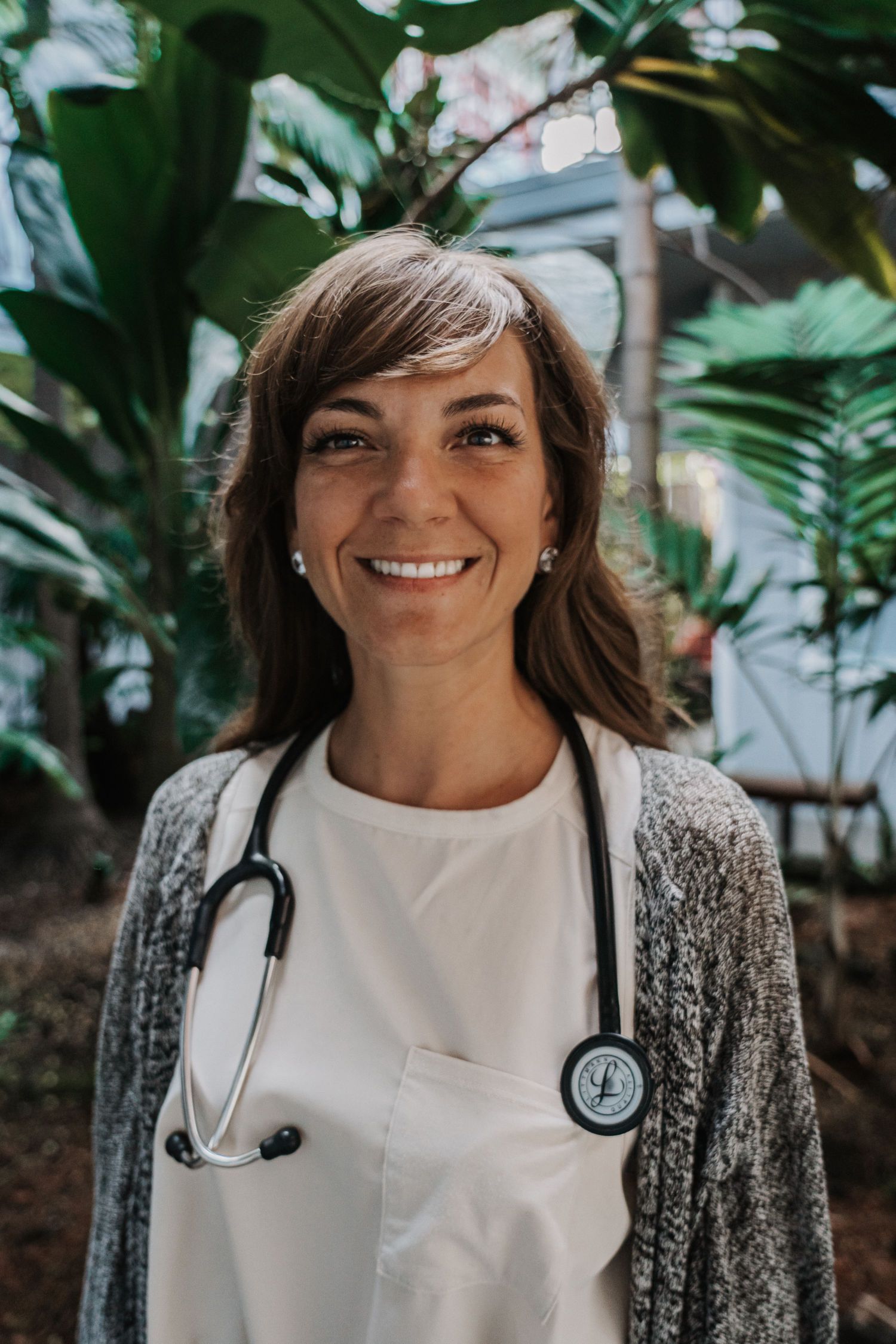 Woman with stethoscope smiles, wearing white scrubs and gray sweater, in a green, natural setting.