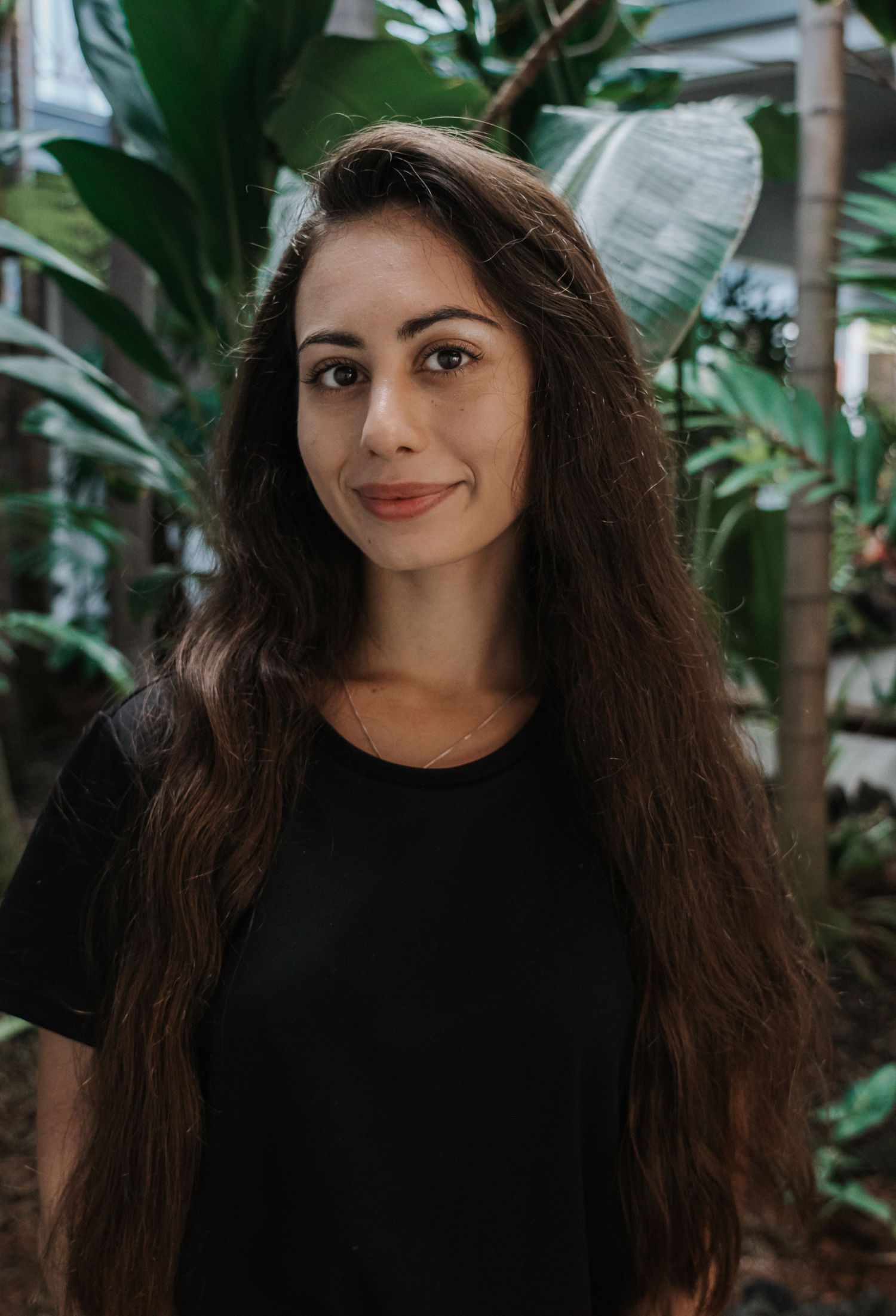 Woman with long brown hair smiling, wearing a black shirt, standing in a garden setting.