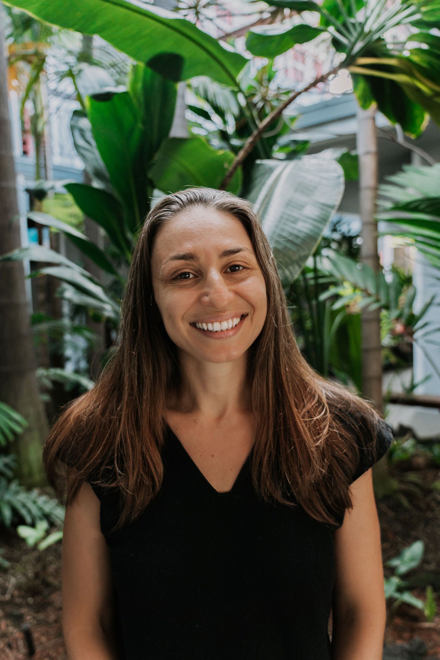 Woman with brown hair smiles, wearing black top in front of lush green plants.