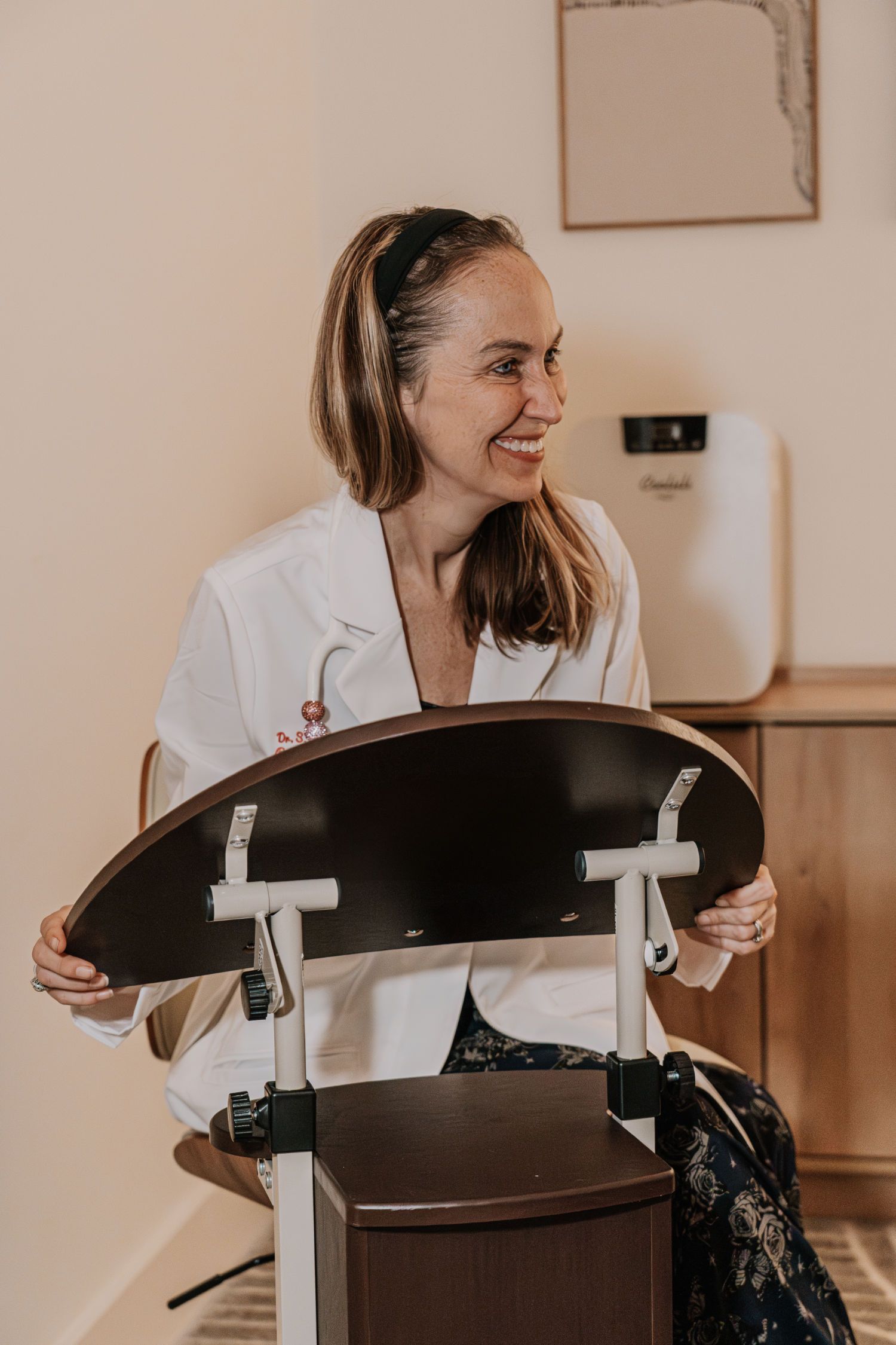 Woman in a lab coat smiles, holding a wheelchair tray in a medical office.