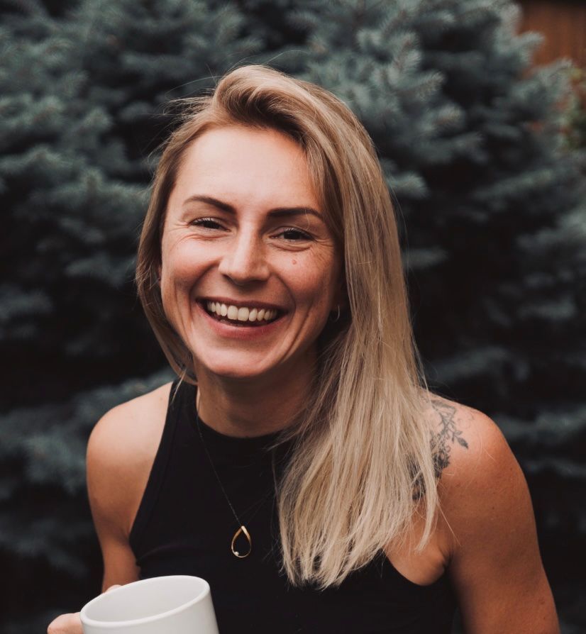Woman with blonde hair, smiling, wearing a black top and gold jewelry, in front of a light blue wall.