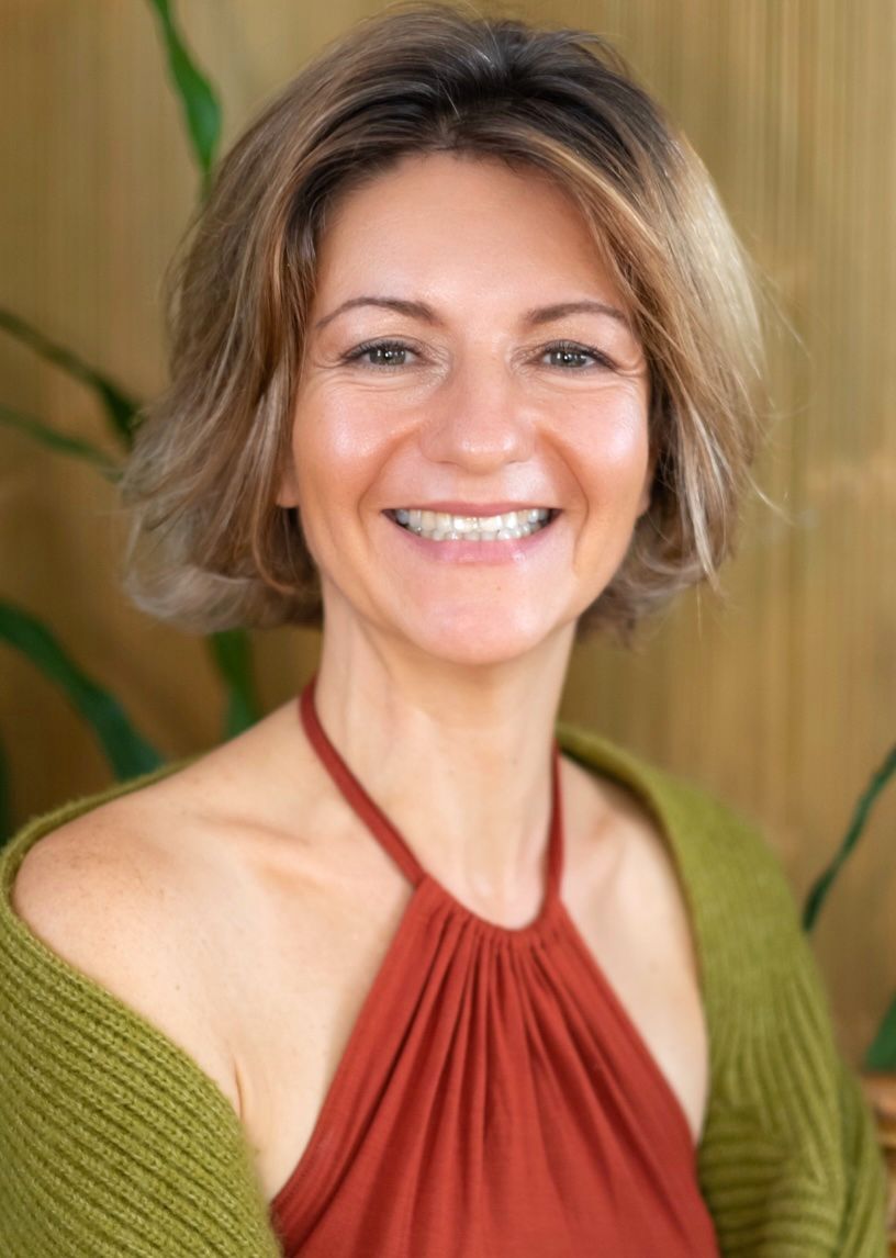 Woman with long brown hair smiles, wearing a green tank top, set against lush green foliage.