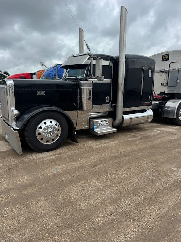 Black Peterbilt semi-truck with chrome accents parked outdoors on dirt under cloudy sky.