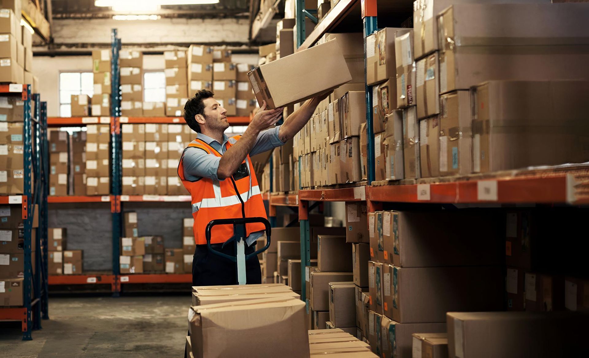 Warehouse worker in orange vest, reaching for a box on a high shelf. Boxes fill shelves.