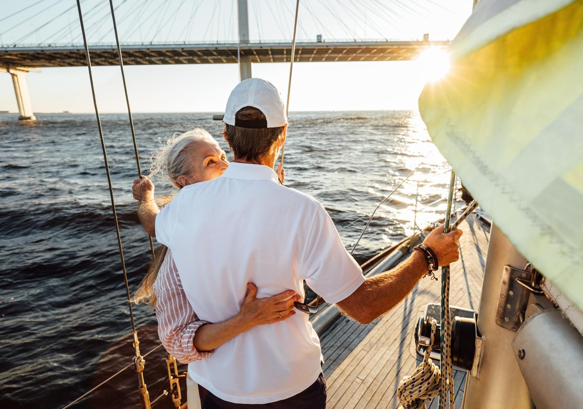 Man embraces woman on a sailboat, sun setting, bridge in the background.