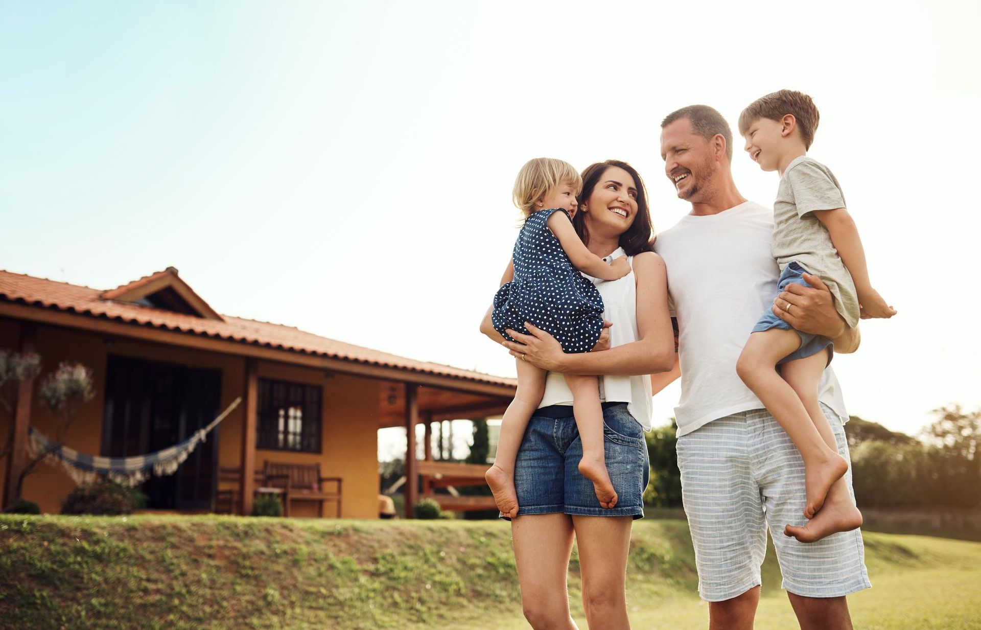 Family of four smiling outside their home. Mom and dad holding children, green lawn, sunny day.
