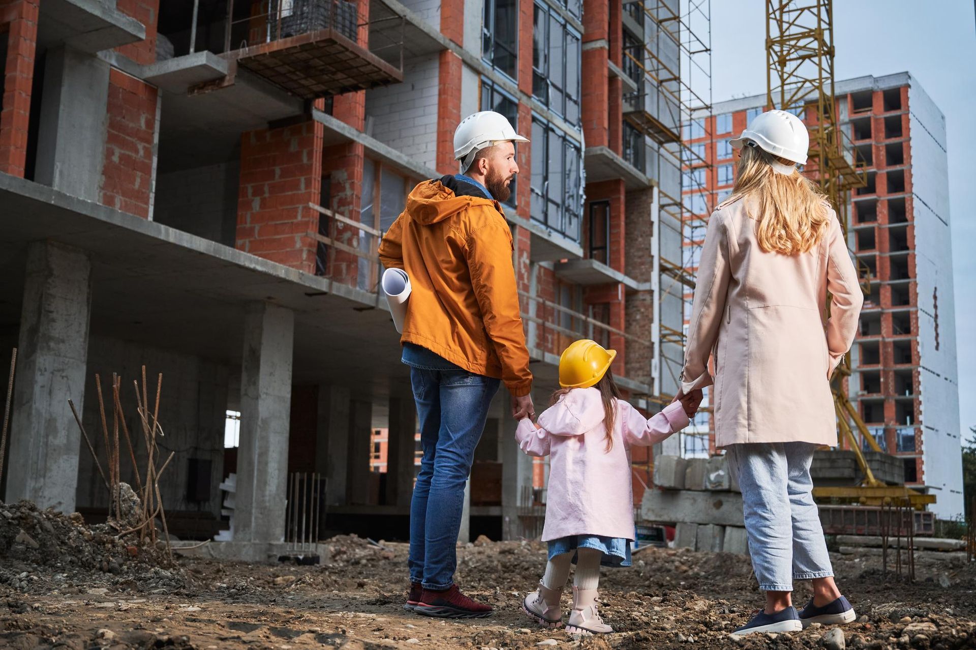 Family in hard hats looking at a building under construction; man holding blueprints.