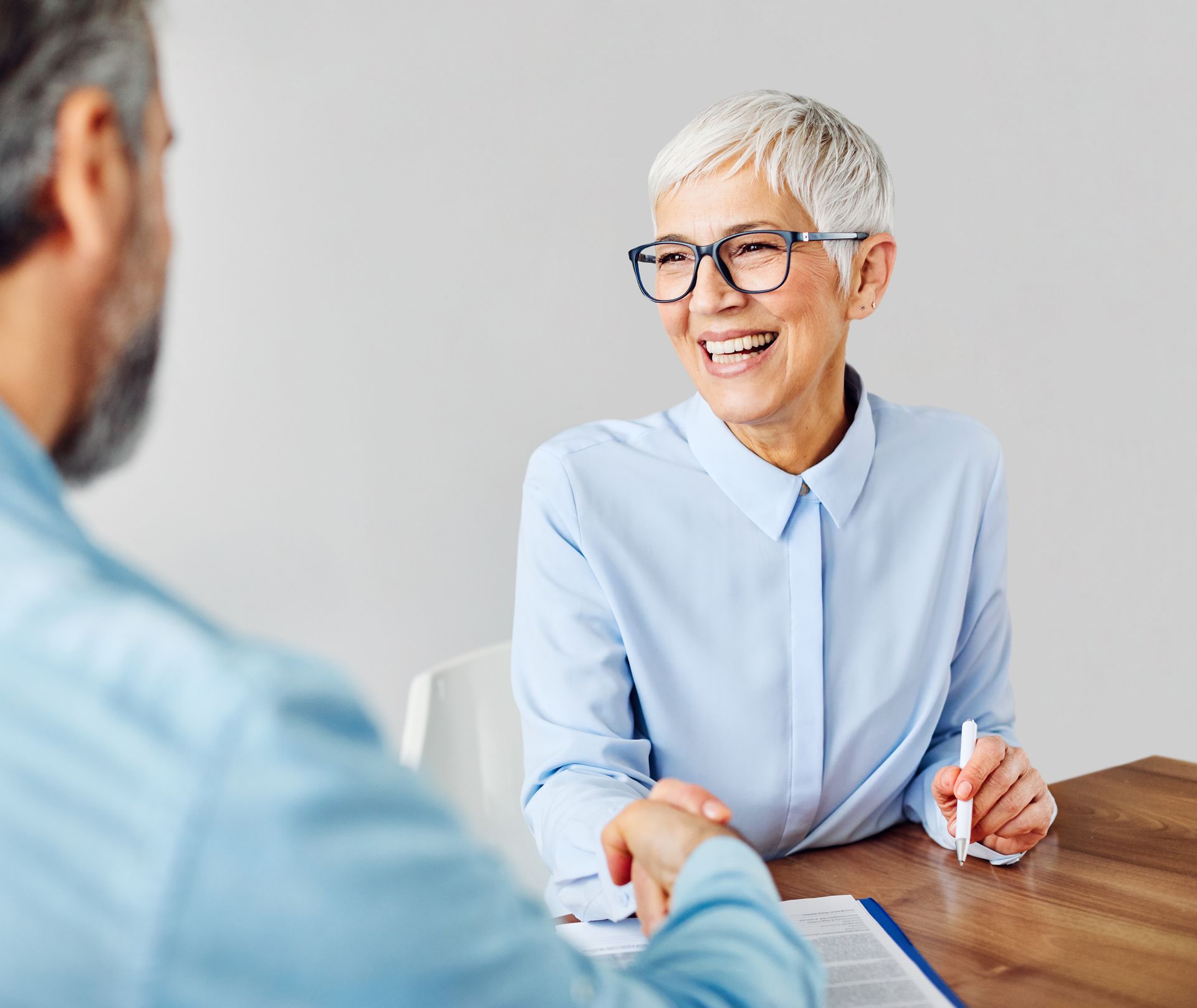 Woman with glasses smiles, shaking hands with a person at a table, possibly a business meeting.