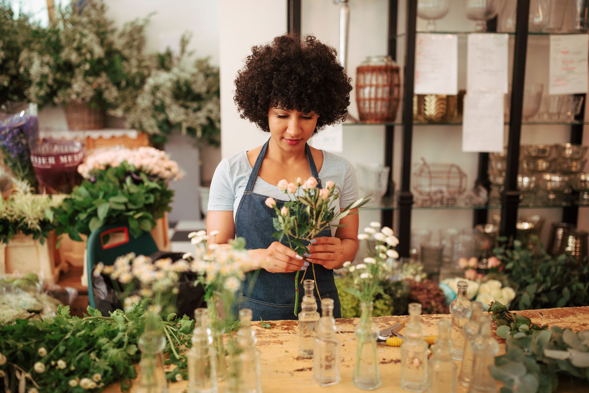 Florist arranging small white flowers in a shop. Woman with afro in blue apron.