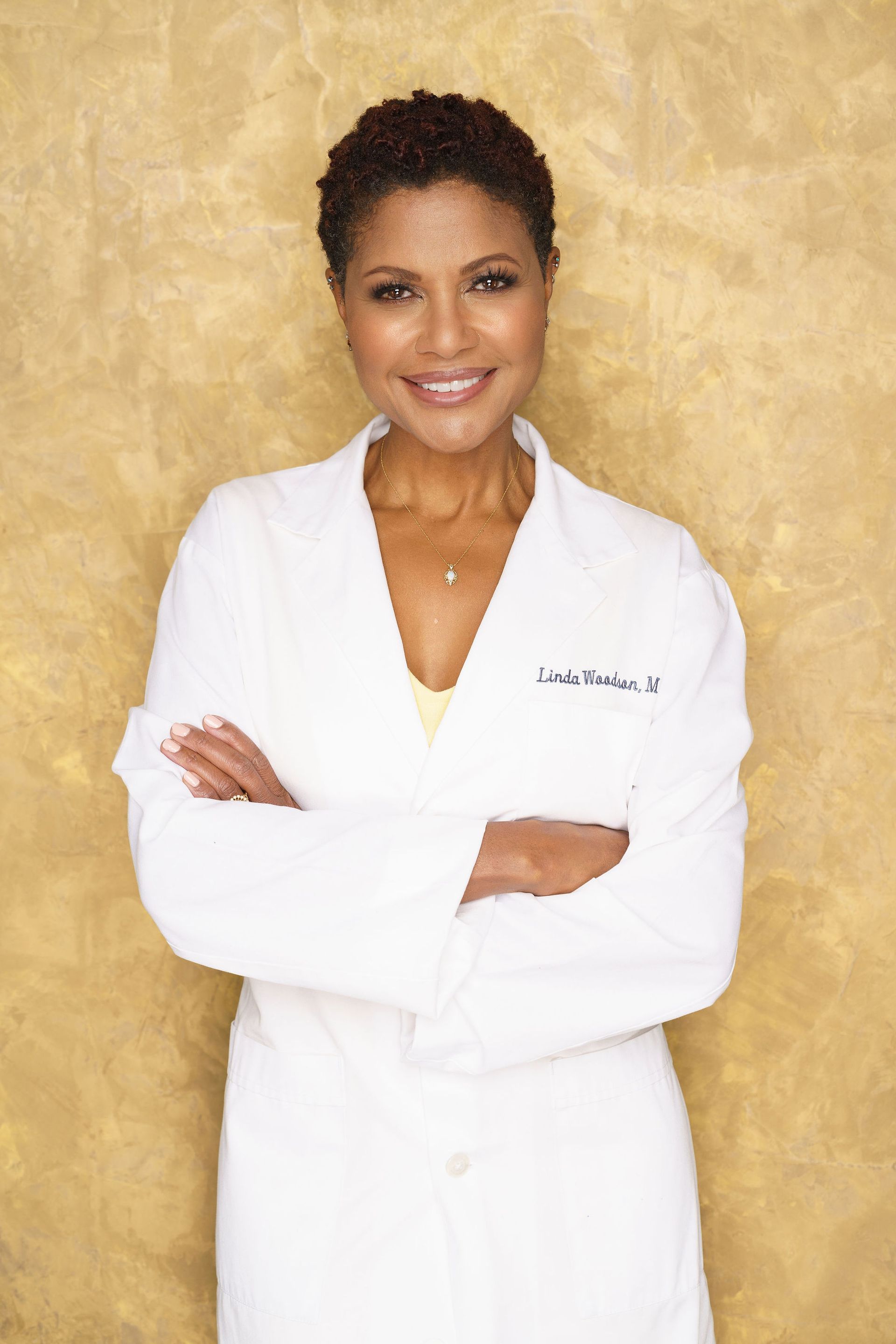 A dermatologist in a white lab coat is smiling with her arms crossed.