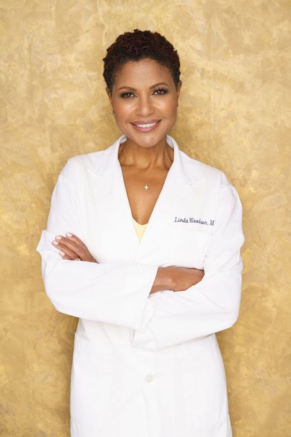 A dermatologist in a white lab coat is smiling with her arms crossed.