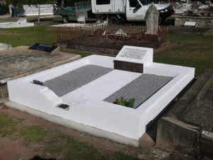 White-painted tomb with two gray gravel sections, small memorial plaque, and greenery in a grassy cemetery.