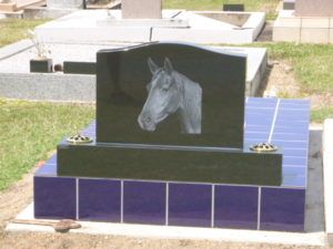 Gravestone with horse head etching on black stone, set on blue tiled base in a cemetery.