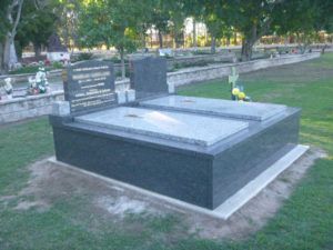 Gray granite double tombstone in a cemetery with trees and grass.