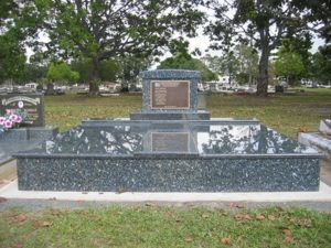 A large granite tombstone with two bronze plaques, set in a grassy cemetery.