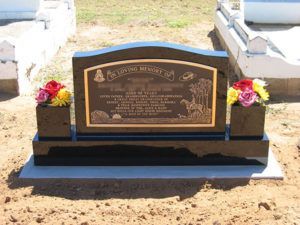 Black granite headstone with bronze plaque and flowers; in a cemetery.
