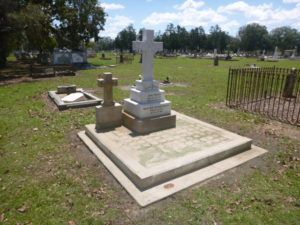 A cemetery scene with a cross-shaped headstone and a smaller cross. Green grass and other headstones are visible.