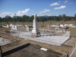 Grave site in a sunny cemetery with white fencing and a large monument. Brown grass and trees in the background.