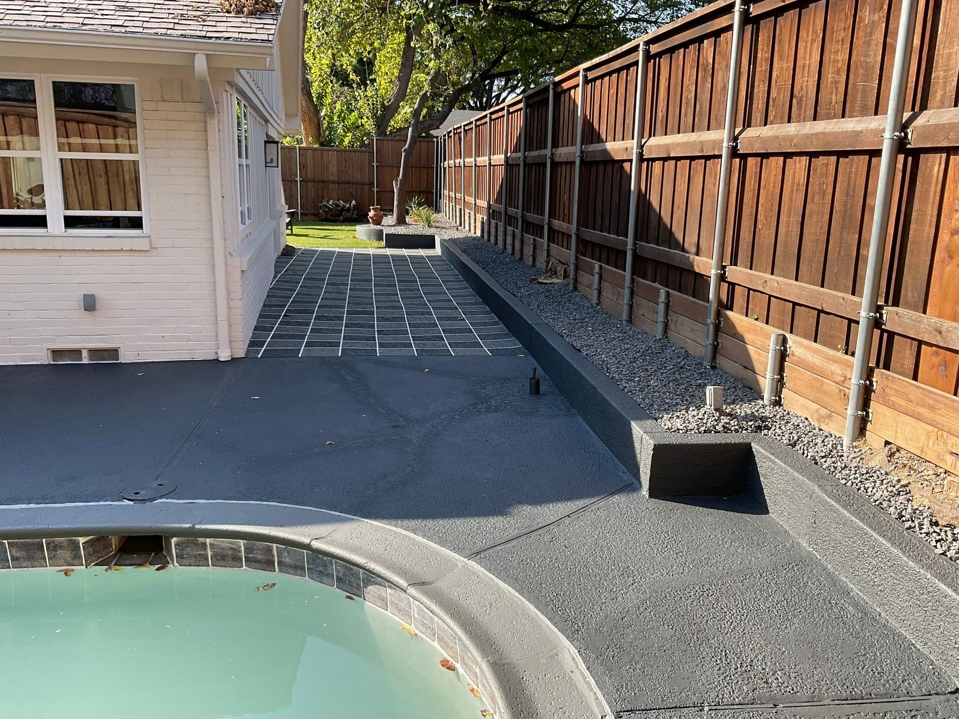 Newly poured concrete patio beside a pool and house, bordering a wooden fence.