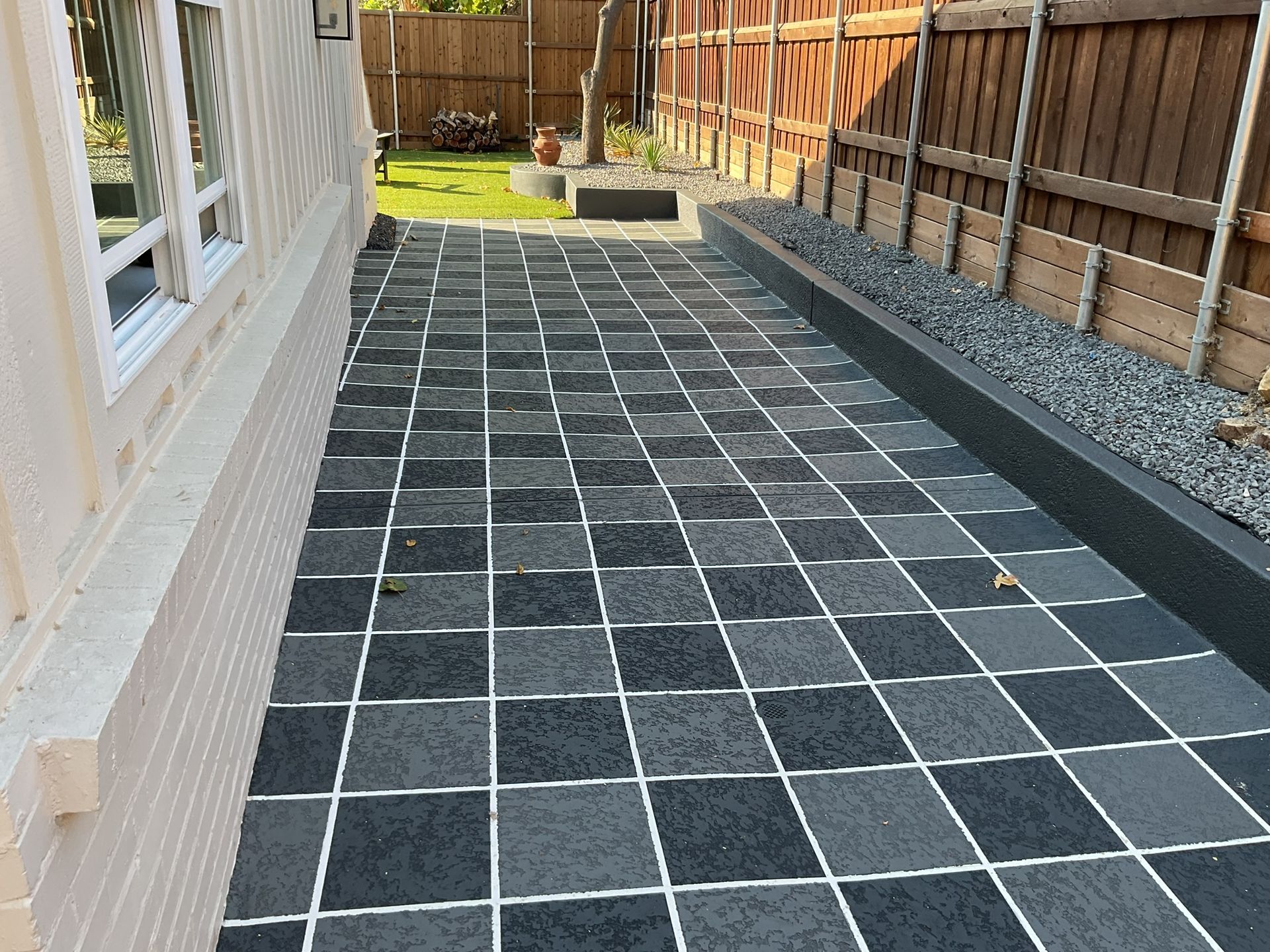 Brick patio with dark gray and light gray square tiles next to a light-colored building and wooden fence.