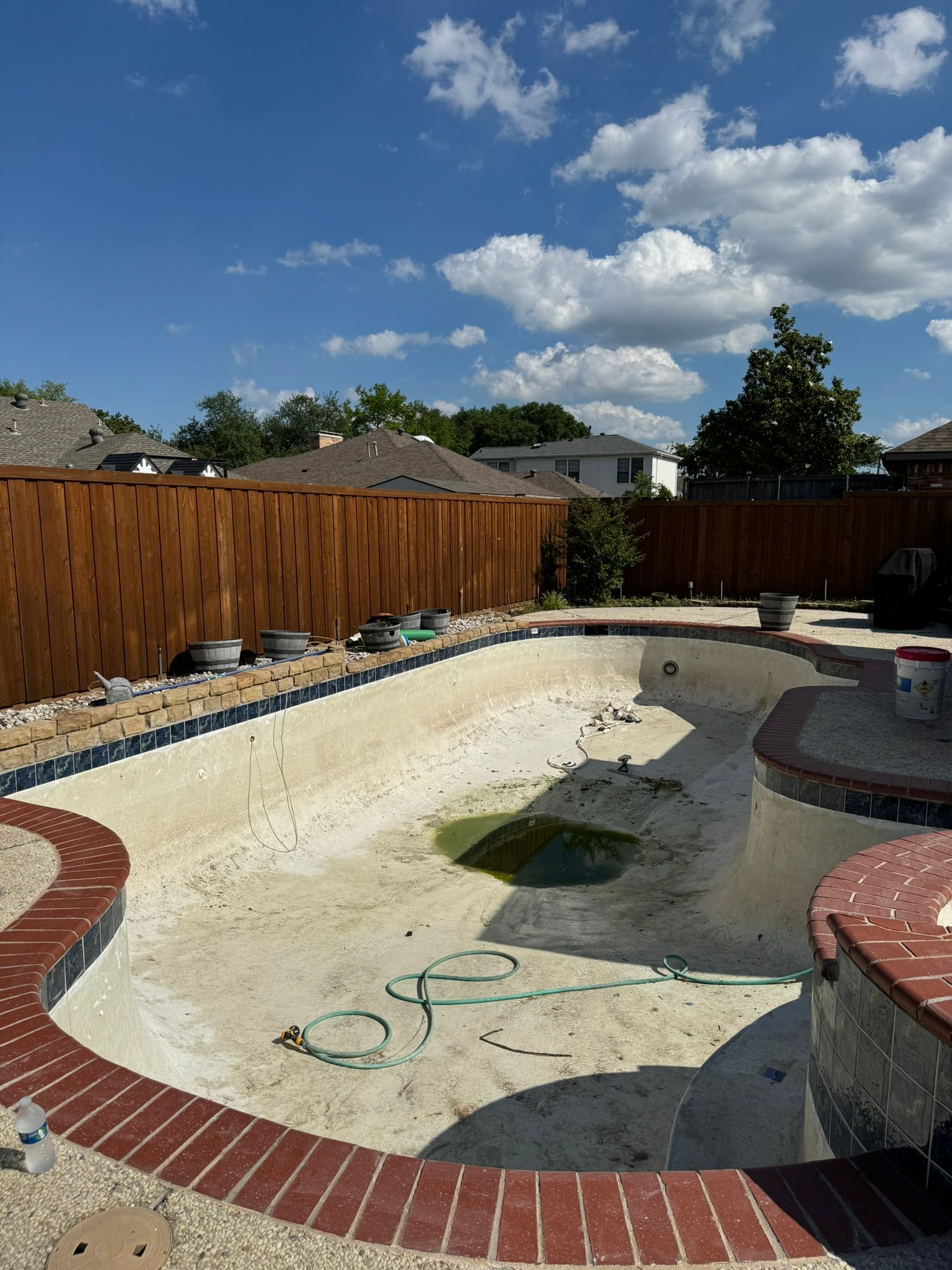 Empty, algae-filled swimming pool with brick trim and wooden fence, under a blue sky with clouds.
