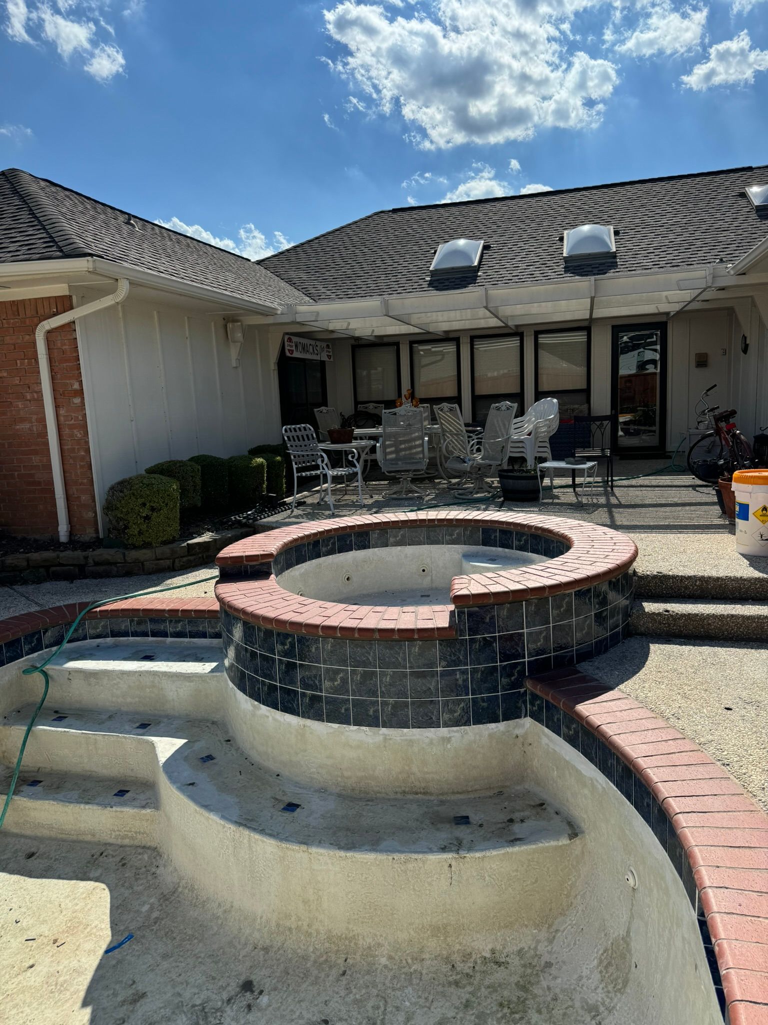 Empty pool with brick edging, patio furniture, and house in the background on a sunny day.